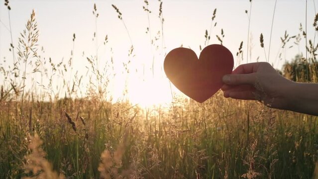 Close up of female hand holding small red heart during beautiful calm sunset or sunrise at summer day. Sunbeams Sunlight. Happy Valentines day, Mothers Day, love nature or life happiness concept