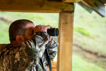 Hunter with his shotgun in middle of nature. Male hunter hunting outdoors, looks into binocular. Hunter men in camouflage clothes with binoculars. Hunter man looks through binoculars.