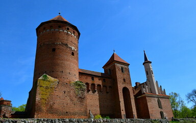 A close up on an old medieval castle made out of red brick, with a  tall tower, angled roof and some addition in the shape of a spire seen on a sunny summer day on a Polish countryside