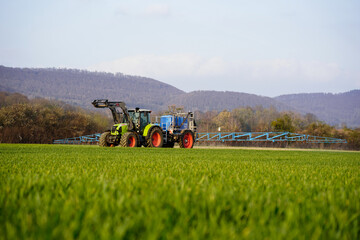 Traktorgespann fährt mit Pflanzenschutzspritze durch jungen Weizenbestand zur Gräserbekämpfung