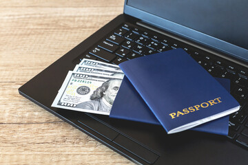 Two blue-covered passports on a black laptop keyboard with U.S. cash dollars on a brown wooden desk. The concept of remote work, study, travel and immigration