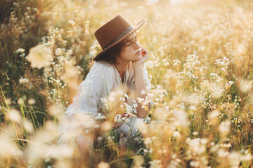 Stylish boho woman in hat sitting among wildflowers in warm sunset light. Summer delight and travel. Young female in rustic linen cloth relaxing in summer meadow. Atmospheric moment