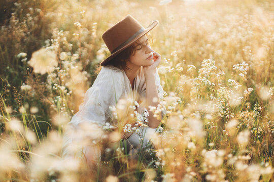 Stylish boho woman in hat sitting among wildflowers in warm sunset light. Summer delight and travel. Young female in rustic linen cloth relaxing in summer meadow. Atmospheric moment