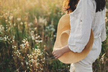 Stylish boho woman walking with straw hat in hand close up among wildflowers in sunset light. Atmospheric moment. Summer travel. Young female in rustic linen cloth relaxing in summer meadow
