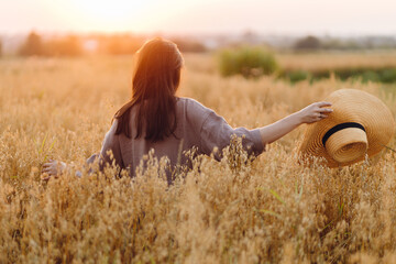 Stylish woman with straw hat standing in oat field in sunset light. Atmospheric tranquil moment. Young female in rustic linen dress relaxing in evening summer countryside. Rural slow life
