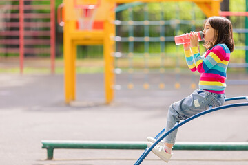 Little girl 5-6 years old, sits on the playground in summer, drinks water and a pink plastic bottle