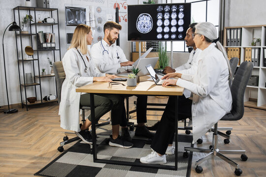 Group of four pensive medical workers looking at patient prescription and considering ways of health treatment. Multicultural coworkers sitting around table with display showing brain CT image.