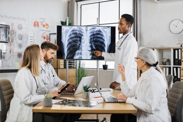 Fototapeta premium African american doctor in lab coat showing x ray scan of lungs on big monitor during conference meeting with international colleagues. Concept of brainstorming, medicine and cooperation.