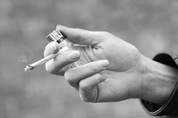 Hand of a young adult women holding a lit hand rolled cigarette and lighter with selective focus. Street life concept in monochrome.