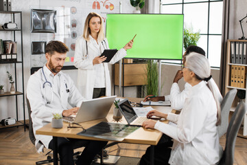 Caucasian female doctor ponting on monitor with green mock up screen while making presentation for international colleagues. Group of medical experts sitting at desk and listening briefing speech.