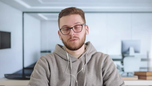 Handsome man in modern earphones sitting at office desh and having video chat, first person view. Concentrated worker listening to colleagues ideas about project strategy.