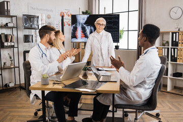 Group of diverse therapists clapping in hands after successful speech of female head doctor at boardroom. Senior woman in lab coat sharing her medical experience with young colleagues on meeting.