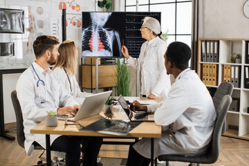 Aged medical worker discussing with multiracial colleagues x ray scan and heartbeat results shown on big digital screen. Team of doctors having briefing at boardroom about severe patient.