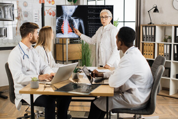 Qualified female doctor leading seminar to three multiracial interns at boardroom. Senior woman standing near digital screen and explaining results of chest x ray scan.