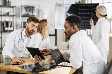 Fototapeta premium Two male doctors sitting at desk and using laptop with tablet while female specialists examining x ray scan on digital monitor. Group of international medical workers having meeting at boardroom .