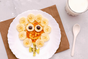 Funny chamomile flower face shape snack from pancake,bananas,kiwi.Cute kids childrens baby's sweet dessert,healthy breakfast,lunch,food art with milk on plate,gray concrete background,top view
