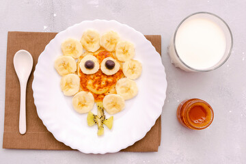 Funny chamomile flower face shape snack from pancake,bananas,kiwi.Cute kids childrens baby's sweet dessert,healthy breakfast,lunch,food art with milk,honey on plate,gray concrete background,top view