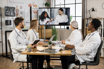 Fototapeta premium Team of male and female doctors having online seminar for international colleague while sitting together at clinic office. Medical workers using digital screen during video conference.