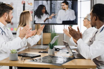 Fototapeta premium Group of male and female doctors cheering with applause their multicultural colleagues during online medical presentation at clinic. Focus on modern digital screen.
