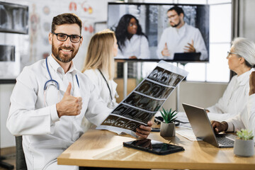 Male orthopedist holding patient x ray result and looking at camera with calm face. Caucasian medical worker having distant healthcare seminar with multiracial coworkers in clinic meeting room.