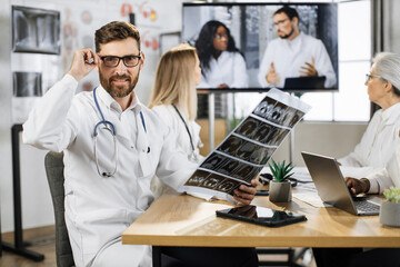 Male orthopedist holding patient x ray result and looking at camera with calm face. Caucasian medical worker having distant healthcare seminar with multiracial coworkers in clinic meeting room.