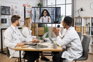 Team of multicultural doctors in white lab coats clapping hands during online video conference after successful speech of excited female scientist. Focus on african american woman on monitor screen.