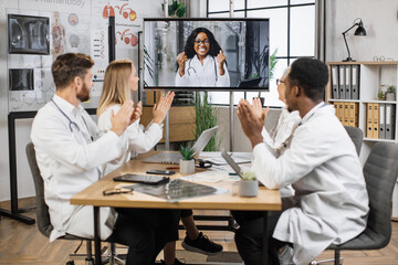Team of multicultural doctors in white lab coats clapping hands during online video conference after successful speech of excited female scientist. Focus on african american woman on monitor screen.