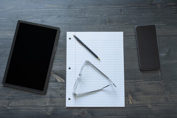 Desk top from top view looking at tablet, paper, pen, phone, and pair of glasses.