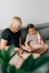 Granny and preschool girl using tablet together at home on the sofa, family togetherness time 
