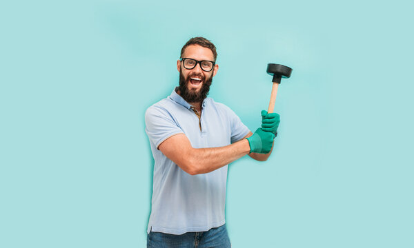 Young handsome man wearing plumber uniform holding toilet plunger looks happy. Professional cleaning of clogged pipes. Studio shot on blue background. Funny promotion poster