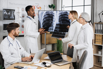 Caucasian female doctor holding x ray scan and talking with multiethnic colleagues while standing near monitor with patient result. Foure medical therapists cooperating for making diagnosis.