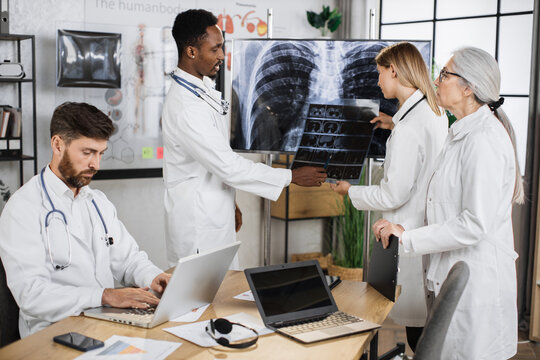 Caucasian female doctor holding x ray scan and talking with multiethnic colleagues while standing near monitor with patient result. Foure medical therapists cooperating for making diagnosis.
