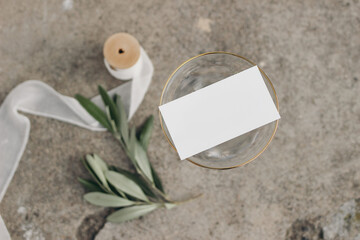 Blank business card mockup on champagne glass with golden rim. Blurred concrete ground with olive...