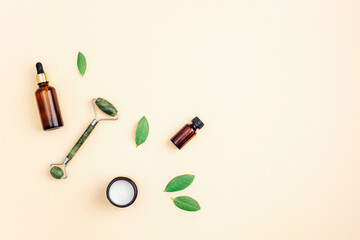 Cream in a jar, serum bottle and jade facial roller on neutral beige background with green leaves, top view. Flat lay, copy space