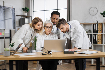 Multiethnic physicians discussing x ray scan while standing around senior female doctor in hospital. Competent elder woman sitting at desk in front of laptop and giving consultation for collegues.