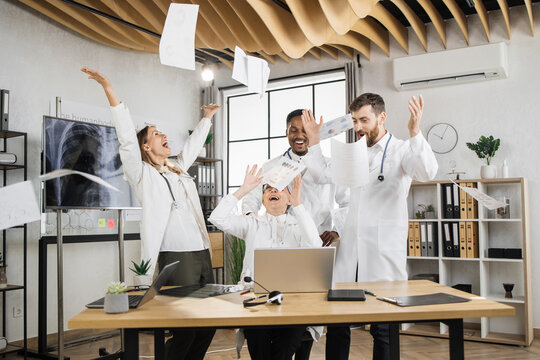 Excited multiracial scientists in white medical uniform laughing and throwing documents in air during laboratory meeting. Success teamwork and healthcare achievement concept.