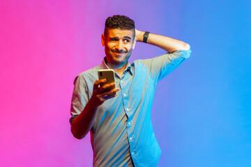 Portrait of man in shirt scratching head holding smartphone in hand, contemplating about software updating, choosing suitable tariffs. Indoor studio shot isolated on colorful neon light background.