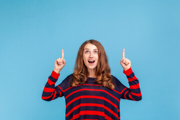 Amazed woman wearing striped casual style sweater, looking astonished and pointing up copy space,...