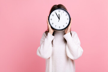 Unknown female hiding her face behind big wall clock, time management, schedule, meeting appointment, wearing white casual style sweater. Indoor studio shot isolated on pink background.