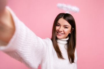 Angelic female with nimb over her head making point of view photo, having happy expression, POV, wearing white casual style sweater. Indoor studio shot isolated on pink background.
