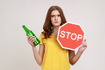 Stop drinking alcohol. Serious teenager girl in yellow T-shirt holding red stop sign and alcohol...