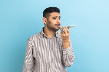 Portrait of self confident young businessman man holding cell phone in hand and talking, leaving voice mail, wearing striped shirt. Indoor studio shot isolated on blue background.