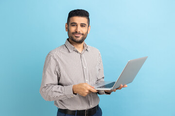 Smiling man freelancer with beard standing and holding laptop, satisfied with teleworking, likes...