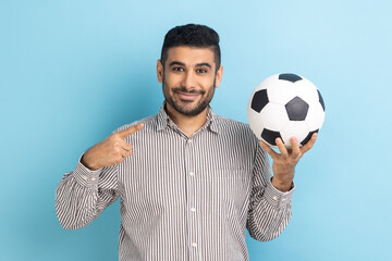 Portrait of handsome businessman pointing finger at soccer ball on his hand with smiling positive expression, wearing striped shirt. Indoor studio shot isolated on blue background.