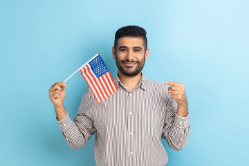 Portrait of happy positive man with beard standing and pointing at american flag, celebrating...