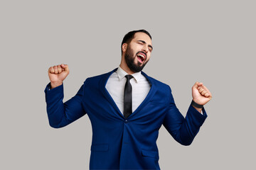 Portrait of sleepy tired exhausted bearded businessman standing and yawning with closed eyes and raised arms, wearing official style suit. Indoor studio shot isolated on gray background.