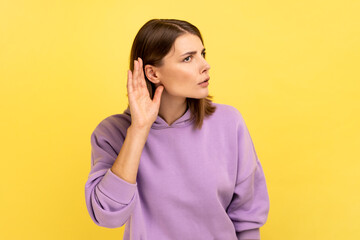 Fototapeta Portrait of pretty woman holding hand near ear, listening attentively with interest private conversation, confidential talk, wearing purple hoodie. Indoor studio shot isolated on yellow background. obraz