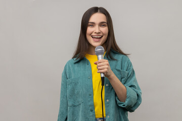 Portrait of positive woman reporter standing with microphone in hands, sing songs with happy expression or telling news, wearing casual style jacket. Indoor studio shot isolated on gray background. - obrazy, fototapety, plakaty
