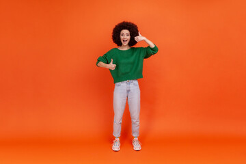 Fototapeta premium Full length portrait of happy friendly woman with Afro hairstyle wearing green casual style sweater standing with thumbs up, expressing positive. Indoor studio shot isolated on orange background.