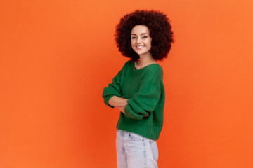 Obraz na płótnie Canvas Side view portrait of smiling woman with Afro hairstyle wearing green casual style sweater standing with folded hands and looking at camera. Indoor studio shot isolated on orange background.